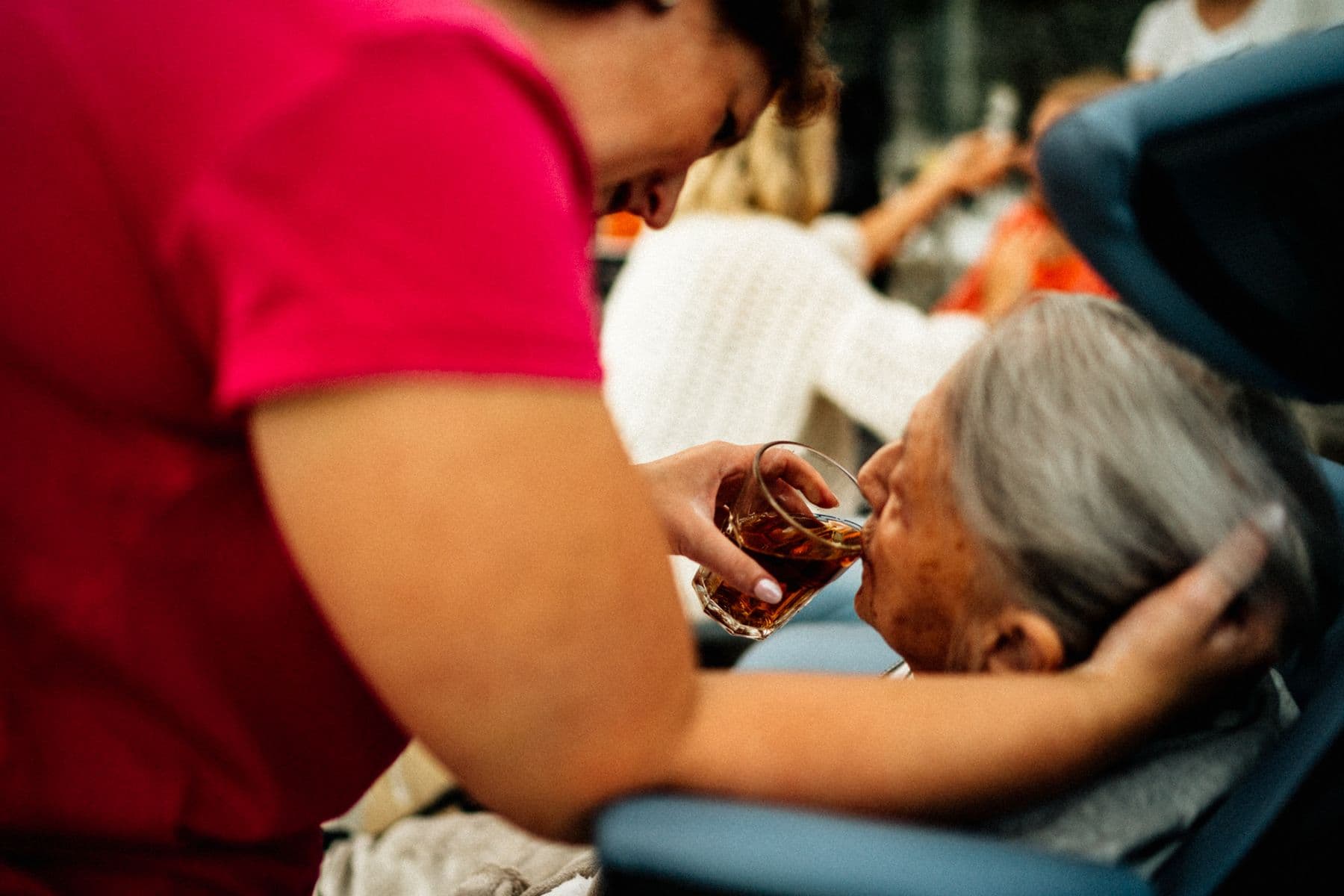 A caregiver gently helps an elderly woman sip tea, close and unhurried